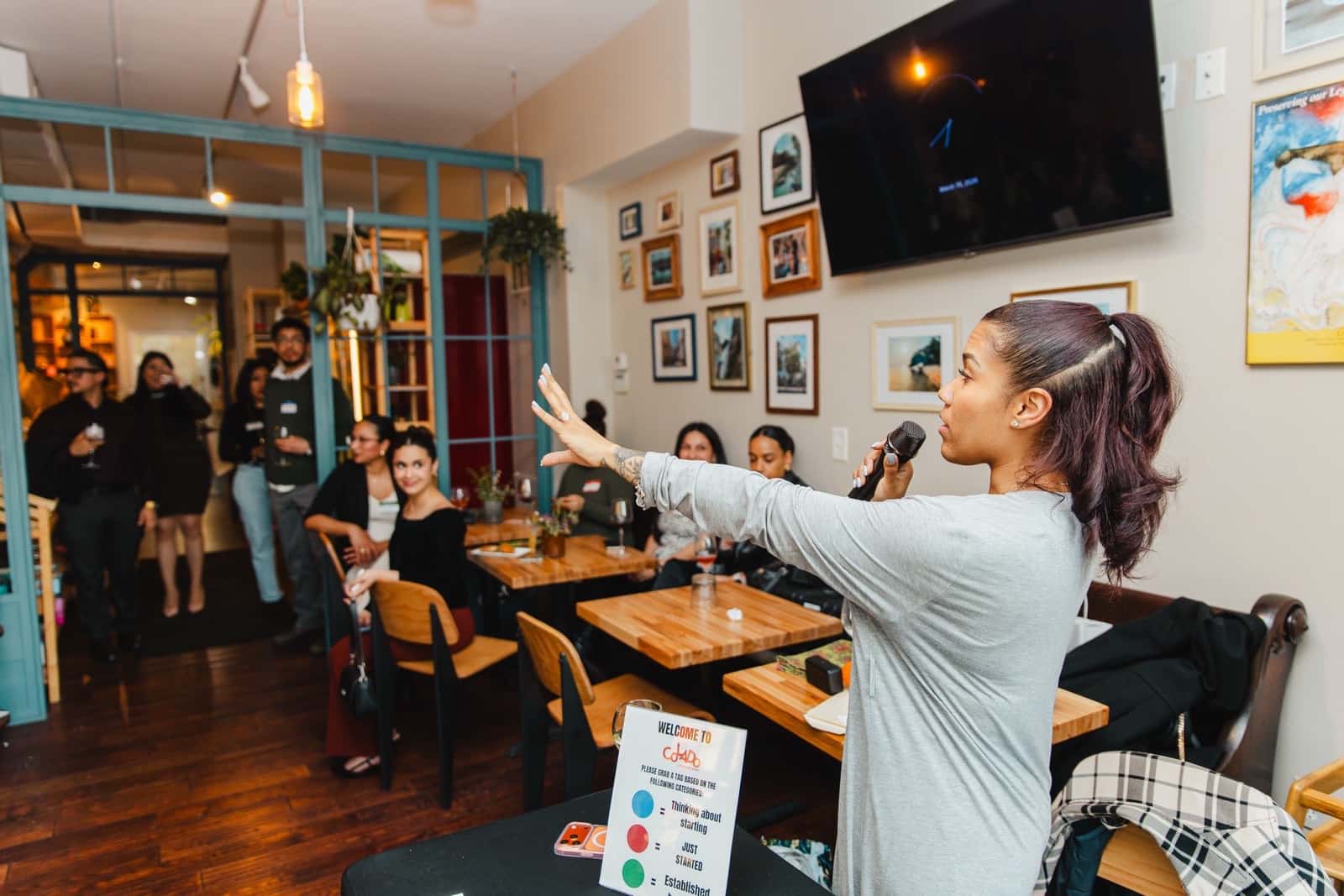 A woman with a microphone talks to the room at Bodega and Vino in Locust Point during Colado Latino Network’s first gathering in March 2026, with guests listening from the wooden tables and the doorway and a Welcome to Colado sign on the front table