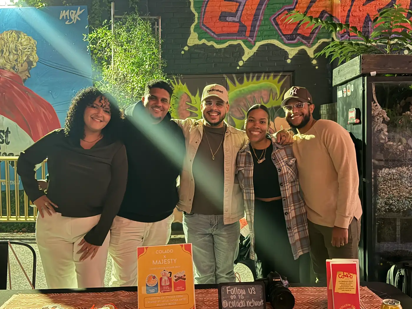 The Colado Latino Network team smiling behind their vendor table at Cultura in Fells Point, Baltimore, with a graffiti mural behind them and the Colado x Majesty sticker flyer, brochures and a Follow us on IG chalkboard on the table