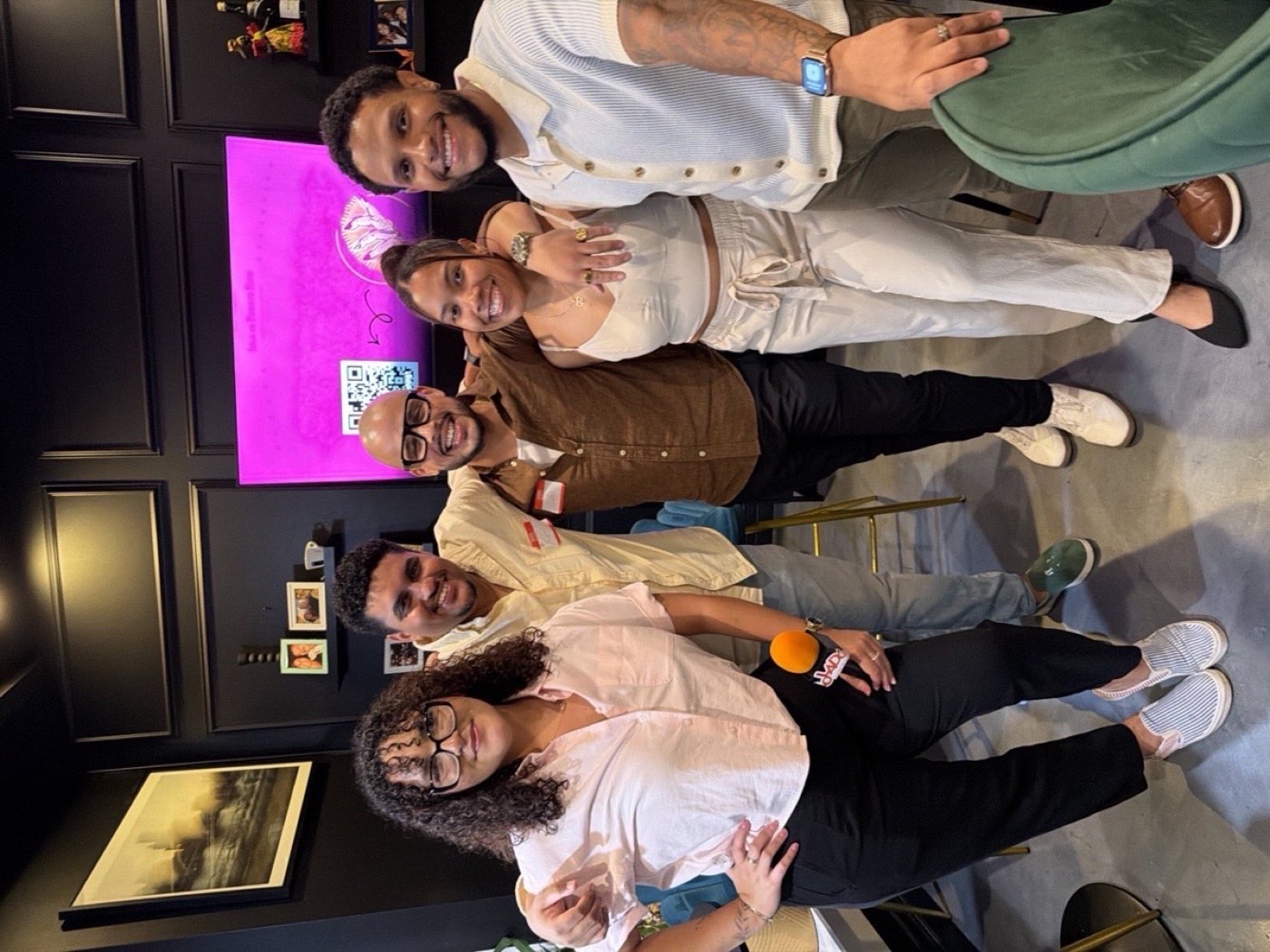 Five guests arm in arm at the Colado Latino Network event at The Empanada Lady in downtown Baltimore, with the Isla de Puerto Rico screen glowing behind them