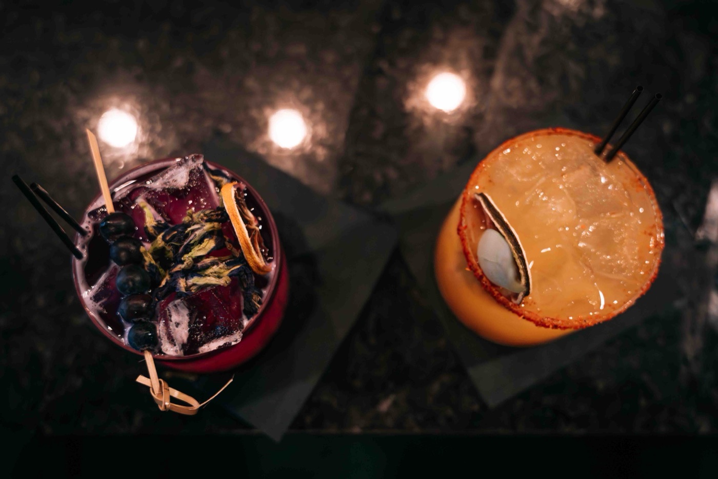 Two craft cocktails from above at The Empanada Lady during the Colado Latino Network gathering, one purple with blueberries and one orange with a chili rim