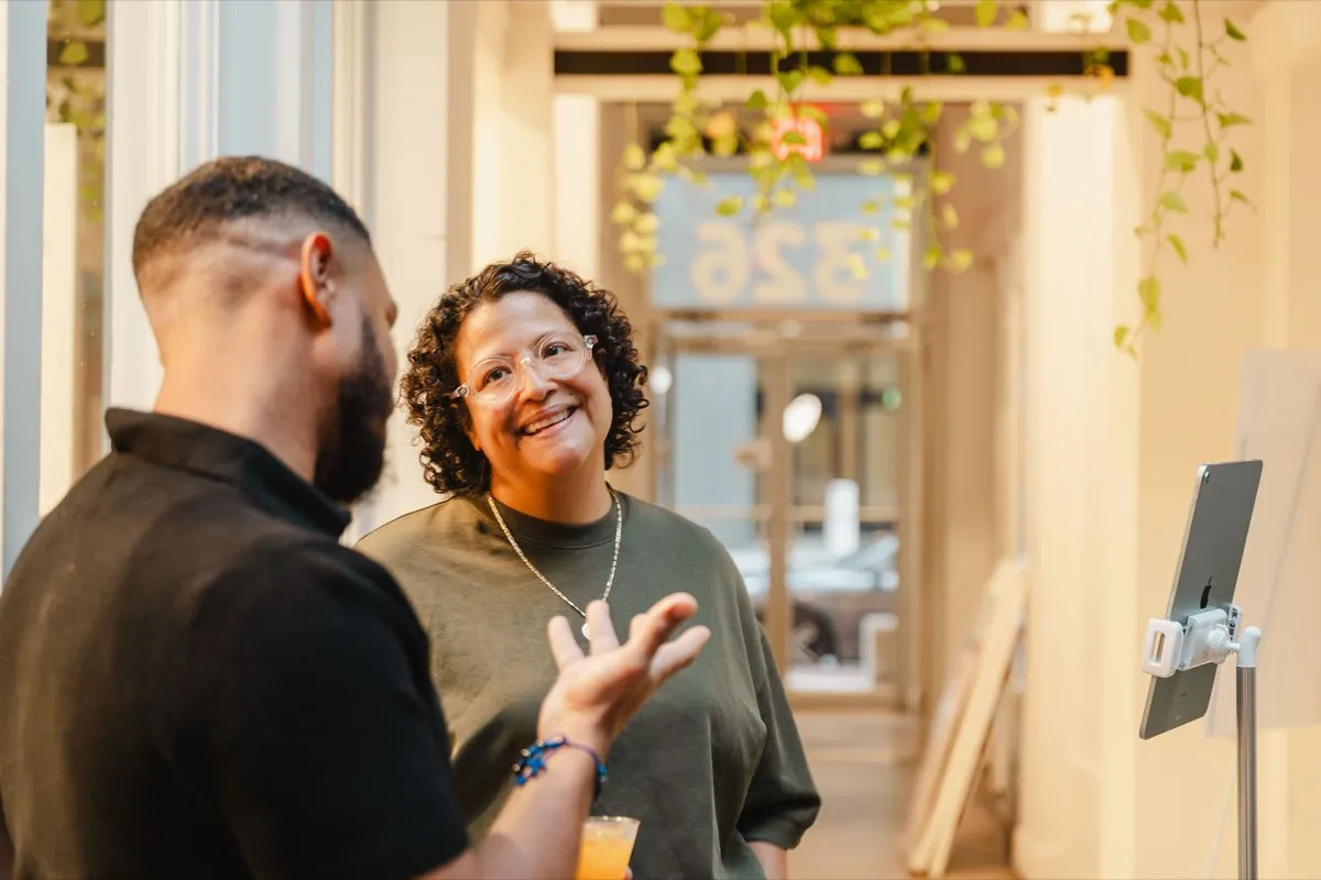Two guests talking near the entrance at a Colado Latino Network gathering in Baltimore, a woman in a green shirt and clear glasses listening while a man gestures with a drink in his hand